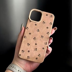 Könnte beinhalten: Eine beige Handyhülle mit floralem Muster. Die Hülle hat einen goldfarbenen Rand um die Kameraöffnung und die Kanten. Kleine, dunkelbraune Blumenmuster sind gleichmäßig auf der Oberfläche verteilt.