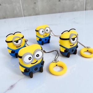 Könnte beinhalten: Gelbe AirPod-Hüllen im Minion-Stil mit blauen Overalls und schwarzen Akzenten. Jede Hülle hat einen passenden gelben Ring mit einem kleinen Gesicht. Die Hüllen sind so gestaltet, dass sie den beliebten Zeichentrickfiguren ähneln.