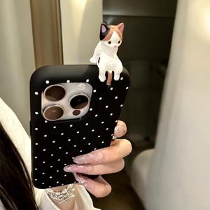 Może przedstawiać: Czarne etui na telefon z białymi kropkami i małą białą figurką kota na górze. Etui na telefon ma wycięcie na obiektyw aparatu.