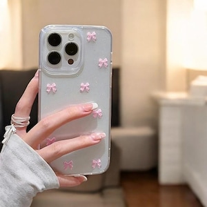 Op de afbeelding: Een transparante telefoonhoes met een glinsterende achtergrond en roze strikjes. De hoes is ontworpen voor een iPhone.