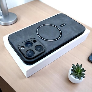 Peut inclure: Une coque de téléphone noire avec une texture en daim gris et un chargeur sans fil intégré. La coque a un renflement de caméra surélevé avec quatre objectifs.