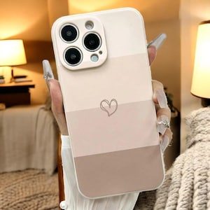 Può includere: Una custodia per telefono bianca con un design a strisce di colore marrone chiaro, beige e marrone scuro. La custodia ha una forma di cuore al centro della striscia beige. La custodia ha una copertura per l'obiettivo della fotocamera rialzata.