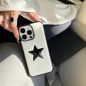 Op de afbeelding: Witte telefoonhoesje met een zwart ster-ontwerp. Het hoesje heeft een zwarte rand en een uitsparing voor de camera.
