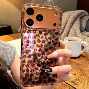 Könnte beinhalten: Transparente Handyhülle mit Leopardenmuster. Die Hülle hat ein goldfarbenes Kameramodul mit drei Linsen und einem Blitz. Die Hülle wird in einer Hand gehalten. Der Hintergrund umfasst eine Kaffeetasse und einen Holztisch.
