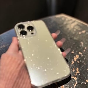 Könnte beinhalten: Eine hellgrüne Handyhülle mit einem glitzernden, gesprenkelten Design. Die Hülle hat eine rechteckige Form mit abgerundeten Ecken und einem Kameraausschnitt. Die Handyhülle wird in einer Hand gehalten.