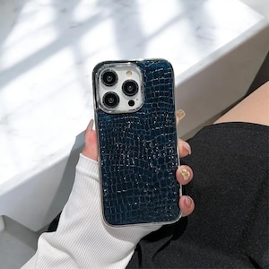 Op de afbeelding: Een blauwe telefoonhoes met een krokodillederen patroon en een zilveren rand. De hoes heeft een transparante camera lens cover met drie zwarte camera lenzen.