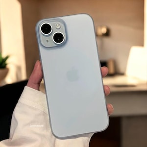 Peut inclure: Une coque de téléphone bleu clair avec une bordure transparente. La coque présente une découpe pour l'appareil photo avec deux lentilles noires et un anneau argenté. Le dos de la coque présente un logo Apple discret.