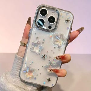 Könnte beinhalten: Transparente Handyhülle mit weißem Glitzerhintergrund und schillernden Sternakzenten. Die Hülle hat einen transparenten Rand und einen Kameraausschnitt. Kleine und große sternförmige Verzierungen sind über die Oberfläche verstreut.