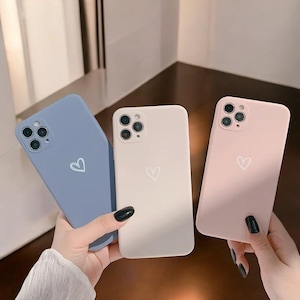 Op de afbeelding: Drie telefoonhoesjes met een hartvormig ontwerp. De hoesjes zijn blauw, wit en roze. Elk hoesje heeft een uitsparing voor de camera en een hartvormig ontwerp.