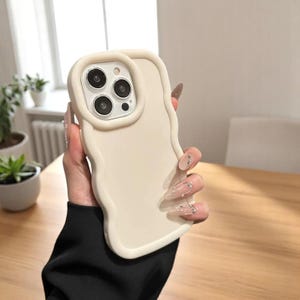 Puede incluir: Funda de teléfono de color crema con un borde ondulado e irregular. La funda tiene un borde elevado alrededor de las lentes de la cámara. La funda del teléfono se sostiene en una mano.