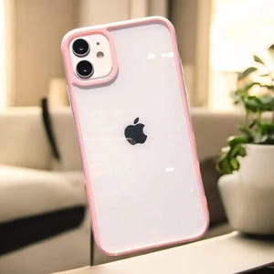 Op de afbeelding: Een transparante telefoonhoes met een roze rand. De hoes heeft een verhoogde camerabult met een heldere lens. Het Apple-logo is zichtbaar op de achterkant van de hoes.