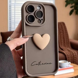 Op de afbeelding: Taupe telefoonhoesje met een verhoogd, beige hartontwerp. De hoes heeft een camera-uitsparing en de naam "Catherine" in cursief schrift. De hoes is gemaakt van glad materiaal.