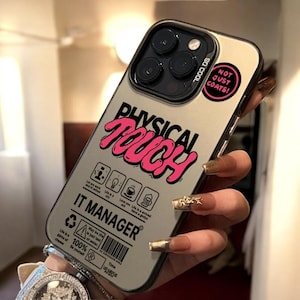 Puede incluir: Una funda de teléfono inteligente con un marco dorado y una parte trasera gris. La funda presenta las palabras "PHYSICAL TOUCH" en rosa, junto con otros textos e iconos. Una pegatina en la funda dice "NOT JUST COATS!"