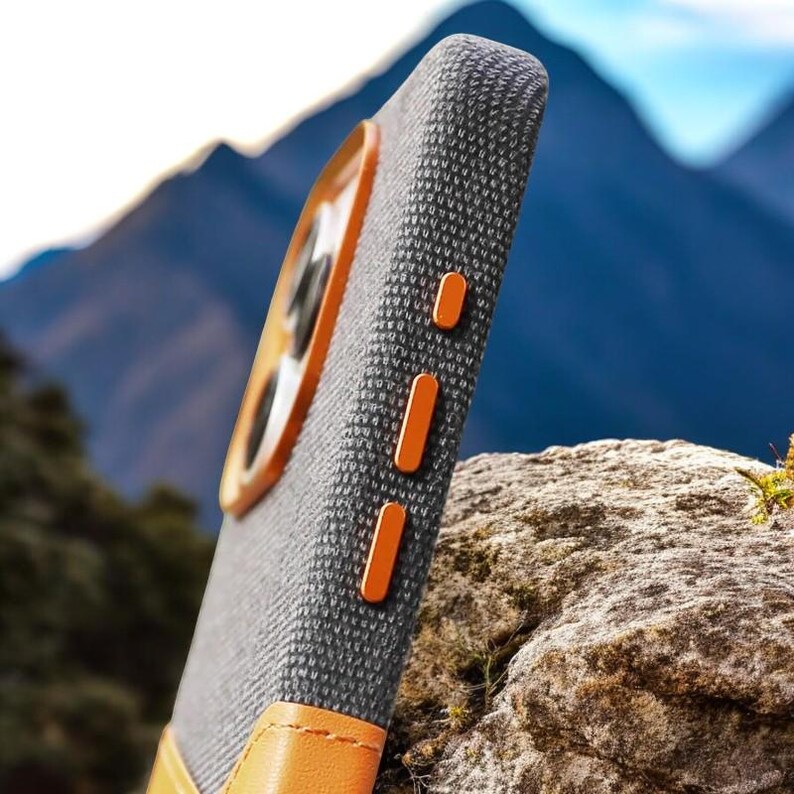 May include: A gray phone case with a brown leather accent and orange buttons. The case is made of a textured fabric and has a raised camera lens.