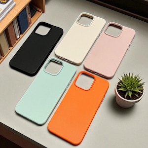 Op de afbeelding: Een verzameling van vijf effen gekleurde telefoonhoesjes in zwart, crème, roze, mintgroen en oranje. De hoesjes zijn gerangschikt op een grijze ondergrond, met een kleine vetplant in een pot op de achtergrond. De hoesjes hebben uitsparingen voor de camera en knoppen.