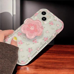 Puede incluir: Una funda transparente para teléfono con un diseño floral rosa y un soporte de anillo en forma de flor rosa con diamantes de imitación. La funda está diseñada para un smartphone con un sistema de cámara dual.