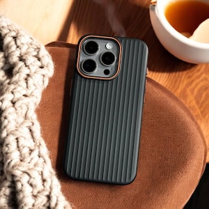 Puede incluir: Funda de teléfono negra con textura acanalada y una carcasa de cámara de color cobre. La funda tiene un diseño moderno y elegante.