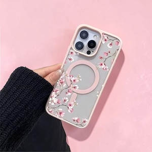 Puede incluir: Funda transparente para teléfono con un diseño floral con flores rosas y blancas. La funda tiene un anillo magnético rosa y un borde rosa. La funda del teléfono se sostiene sobre un fondo rosa.