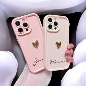 Könnte beinhalten: Zwei Handyhüllen, eine hellrosa und eine cremefarbene, jeweils mit einem goldenen Herzakzent. Die rosa Hülle trägt den Namen "Jess" in Schreibschrift, und die cremefarbene Hülle trägt "Kristin". Beide Hüllen haben einen Kameraschutz.