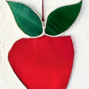 Peut inclure: Une pomme en verre teinté rouge avec des feuilles vertes. La pomme est suspendue à une ficelle.