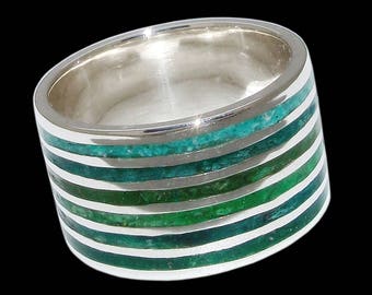 Op de afbeelding: Een zilveren ring met een brede band en meerdere groene strepen. De strepen zijn gemaakt van een materiaal dat eruitziet als turquoise.