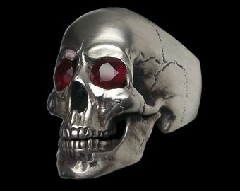 May include: A silver skull ring with red gemstone eyes. The skull is detailed with a textured surface and a toothy grin.