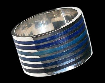 Op de afbeelding: Een brede zilveren ring met een band van afwisselende blauwe en zilveren strepen. De ring heeft een gepolijste afwerking en een modern design. De blauwe strepen variëren in tint, waardoor een gradiënteffect ontstaat. De ring staat tegen een zwarte achtergrond.