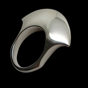 Puede incluir: Un anillo de plata pulida con un diseño escultural único. El anillo presenta una superficie lisa y curvada con una parte superior redondeada y una banda ancha. El diseño del anillo es moderno y minimalista, sobre un fondo negro.