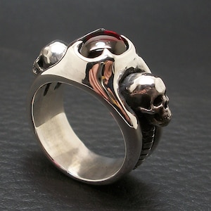 May include: A silver ring with a red gemstone and two skull designs. The ring is made of sterling silver and has a detailed design.