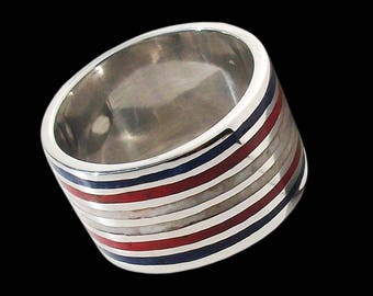 Op de afbeelding: Een brede zilveren ring met afwisselende banden van rode, witte en blauwe emaille. De ring heeft een gepolijste afwerking en een eenvoudig, modern ontwerp. De ring wordt getoond tegen een zwarte achtergrond.