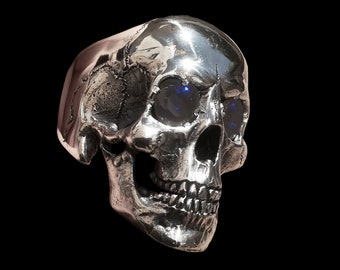 Puede incluir: Un anillo de calavera de plata con ojos de piedras preciosas azules. El anillo está detallado con una superficie texturizada y un diseño de calavera realista.