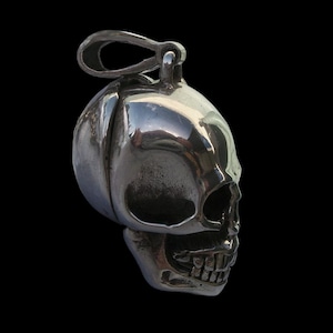 May include: A silver skull pendant with a detailed design. The skull has a hinged jaw and a loop at the top for attaching to a chain.