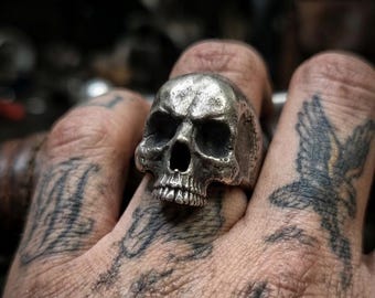 May include: A silver skull ring with detailed facial features, including eye sockets, a nose cavity, and teeth. The ring is worn on a finger, showcasing the intricate design and metallic sheen. The ring is a gothic or biker-style accessory.