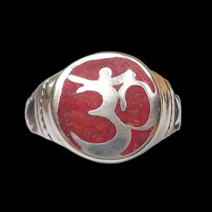 May include: A silver ring with a red inlay featuring the Om symbol. The ring has a polished finish and a detailed design. The Om symbol is a sacred sound and spiritual icon in Hinduism and other religions.