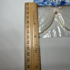May include: White angel wings with gold trim, packaged in a clear plastic bag. The wings are approximately 9 inches in length, as measured by a wooden ruler. The wings are made of a sheer, glittery material.