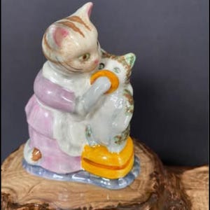 Puede incluir: Figura de cerámica de un gato abrazando a un gato más pequeño. El gato más grande lleva un vestido rosa y un delantal blanco. Los gatos son de color crema con rayas marrones y están sobre una base de madera.