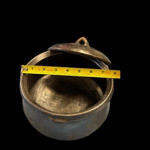 May include: A bronze-colored, round pot with a lid, and a handle. The pot is approximately 7 inches in diameter. The lid has a small loop on top. The pot is set against a black background.