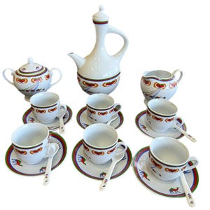 May include: A white ceramic tea set with a decorative design featuring red hearts, camels, and a green, red, and blue border. The set includes a teapot, sugar bowl, creamer, cups, saucers, and spoons.