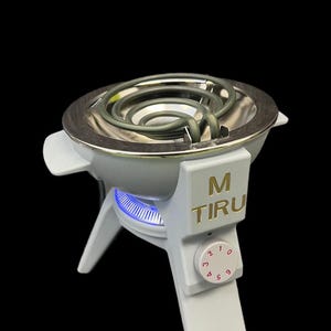 Puede incluir: Una placa caliente eléctrica blanca y plateada con un elemento calefactor circular. La placa caliente tiene una perilla de control con números y el texto "M TIRU" en dorado. El elemento calefactor es de diseño en espiral.