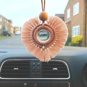 May include: A peach-coloured macrame car hanging ornament featuring a circular ring wrapped in cord with a fringe edge that forms a heart shape. A single smooth wooden bead sits above the ring, attached to a matching cord for hanging purposes.