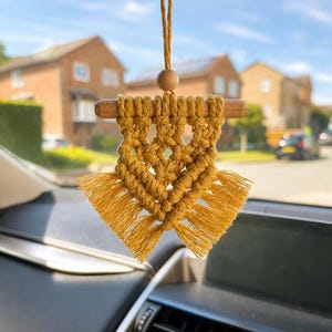 Puede incluir: Un adorno colgante de macramé para coche en color amarillo mostaza con un patrón geométrico anudado suspendido de un pasador de madera. Una cuenta de madera se sitúa sobre el pasador en un cordón a juego y el borde inferior tiene flecos.
