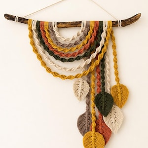 May include: A macrame wall hanging suspended from a dark wooden branch, featuring multiple arched layers of twisted cord in mustard yellow, forest green, terracotta, and cream. Several knotted cord leaves in matching colours hang from the ends.