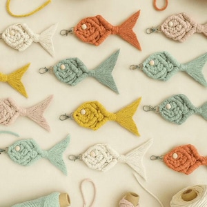 May include: A collection of handmade macrame fish keychains in various colors, including orange, yellow, and blue-green. Each fish has a small wooden bead eye and a metal clasp. Spools of yarn in matching colors are also visible.