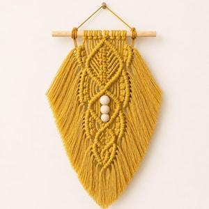 May include: A mustard yellow macrame wall hanging featuring intricate knotted patterns in a leaf-like shape, suspended from a light-coloured wooden dowel. The design includes three smooth, round, light-coloured wooden beads centred vertically.