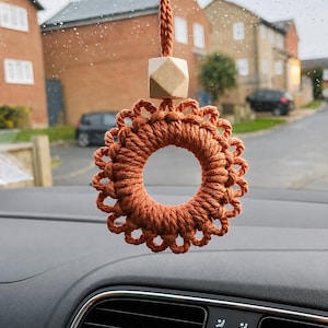 May include: A rust-coloured macrame car charm featuring a circular ring design with a decorative loop border. The ornament is suspended by a matching cord and includes a single, light-coloured, geometric wooden bead positioned above the ring.