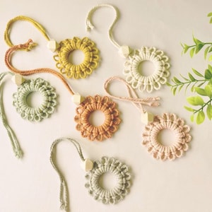 Könnte beinhalten: Eine Sammlung handgefertigter, kreisförmiger Makramee-Ornamente in verschiedenen Pastellfarben, darunter Gelb, Grün, Pfirsich und Rosa. Jedes Ornament hat eine Holzperle und eine Schlaufe zum Aufhängen. Die Ornamente sind auf einer weißen Oberfläche angeordnet.
