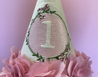 May include: A white fabric party hat with a pink circle and the number "1" embroidered in white. The hat is decorated with pink fabric flowers.
