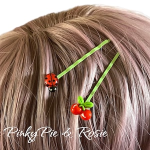 Op de afbeelding: Twee groene haarclips met een lieveheersbeestje en een kersendesign. Het lieveheersbeestje is rood met zwarte stippen en de kers is rood met een groene steel en blad.
