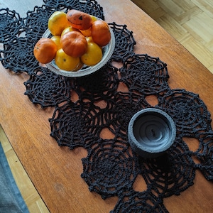 May include: A black, crocheted table runner with a spiderweb design. A silver bowl holds oranges and a carved pumpkin. A gray candle holder with a black candle sits on the table. The table is made of wood.