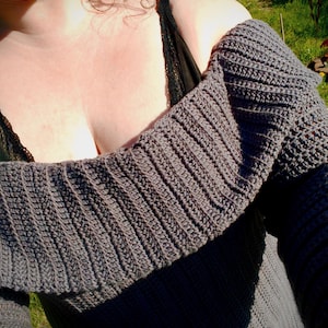 Puede incluir: Un suéter de crochet gris, con hombros descubiertos. El suéter tiene una textura acanalada en las mangas y un cuello ancho y doblado. La prenda está hecha de un material suave, posiblemente lana.